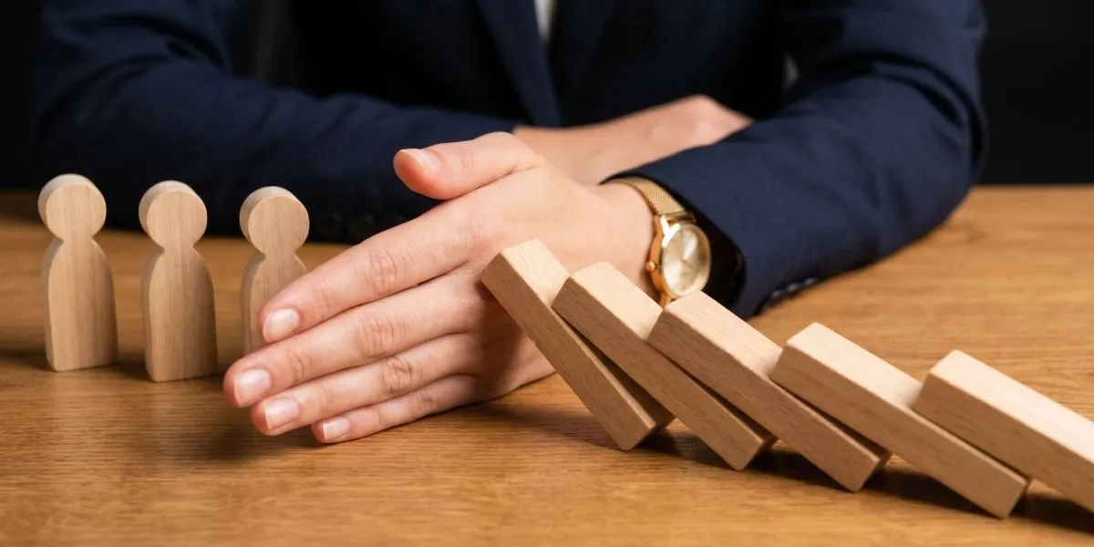 Risikoanalyse: Nahaufnahme der Hand einer Angestellten, sie sitzt an einem Schreibtisch und schützt mit ihrer aufgestellten Hand eine Reihe geraderstehender Dominoklötze, während von der anderen Seite schräg umfallende Dominoklötze zur Seite in Richtung Hand kippen.
