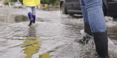 Drainagebeton: Personen gehen mit Gummistiefeln durch eine überflutete Straße, während ein Auto durch das Wasser fährt. Eine Person trägt einen gelben Regenmantel.