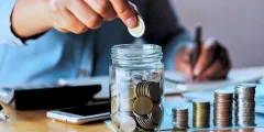 Person legt Geldmünze in Glas mit weiterem Bargeld und notiert sich etwas, daneben ein Taschenrechner und weitere Geldmünzenstapel auf einem Schreibtisch