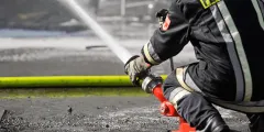 Löschwasserrückhaltung: Ein Feuerwehrmann in schwarzer Schutzkleidung richtet einen Wasserstrahl aus einem roten Hydrantenanschluss auf einen Einsatzort; im Vordergrund verläuft ein gelber Feuerwehrschlauch.