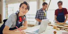 Auszubildende Person in Tischlerei arbeitet an Holzverarbeitungsfabrik, vor ihr ein Laptop und ein Notizheftt, im Hintergrund zwei Kollegen
