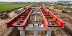 Vergabebeschleunigungsgesetz: Baustelle einer Brücke mit roten Stahlträgern auf Betonpfeilern; drei Bauarbeiter stehen auf einem Brückenabschnitt, während im Hintergrund Bagger, Lastwagen und weitere Baumaschinen arbeiten.